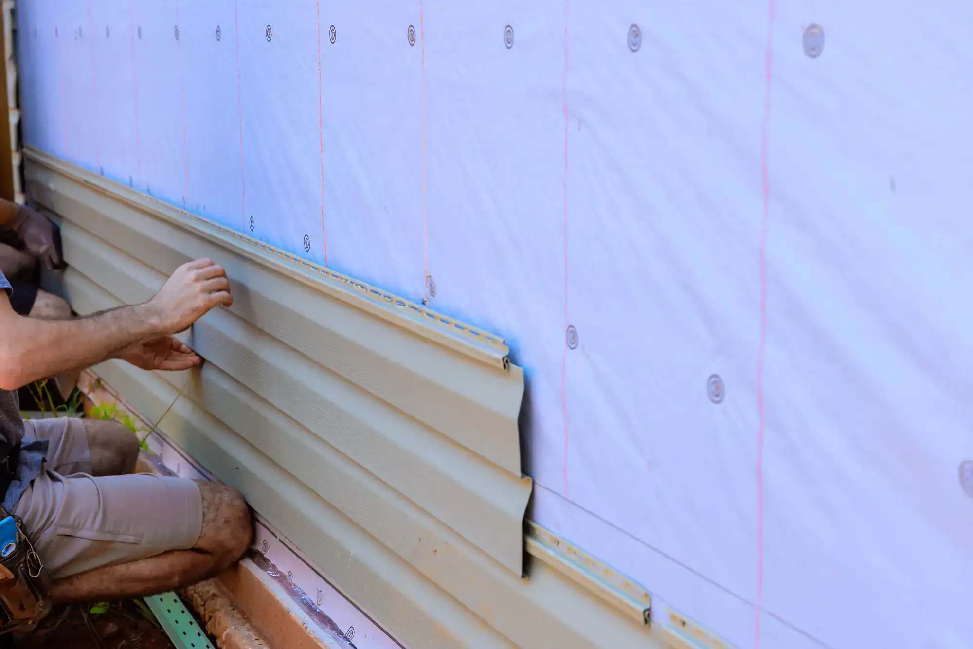 A person installs beige vinyl siding panels on the exterior wall of a house, which is covered with a white weather-resistant barrier.