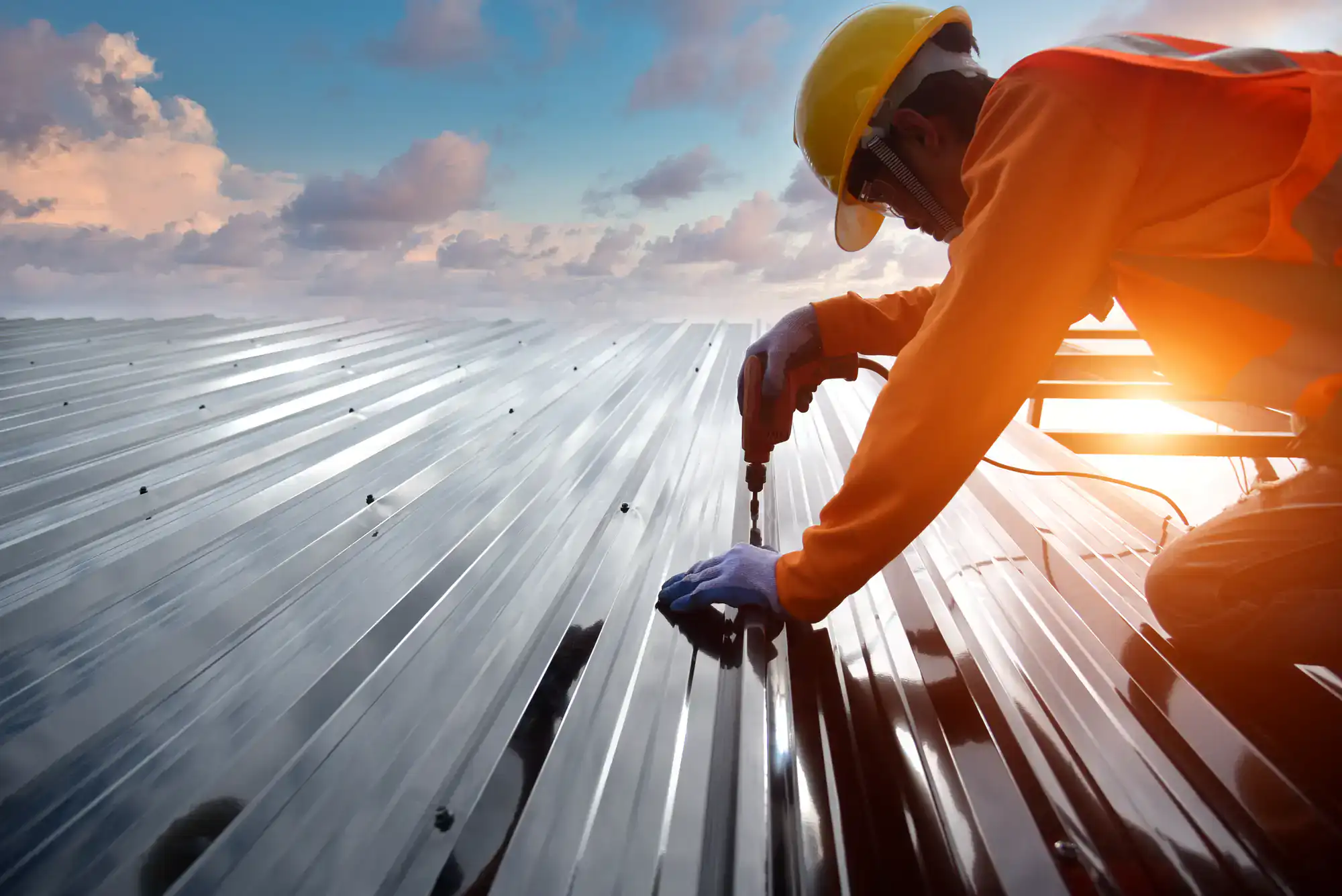 A construction worker wearing a yellow helmet, orange vest, and gloves uses a power drill to secure a metal sheet roof at sunset, with clouds in the sky.