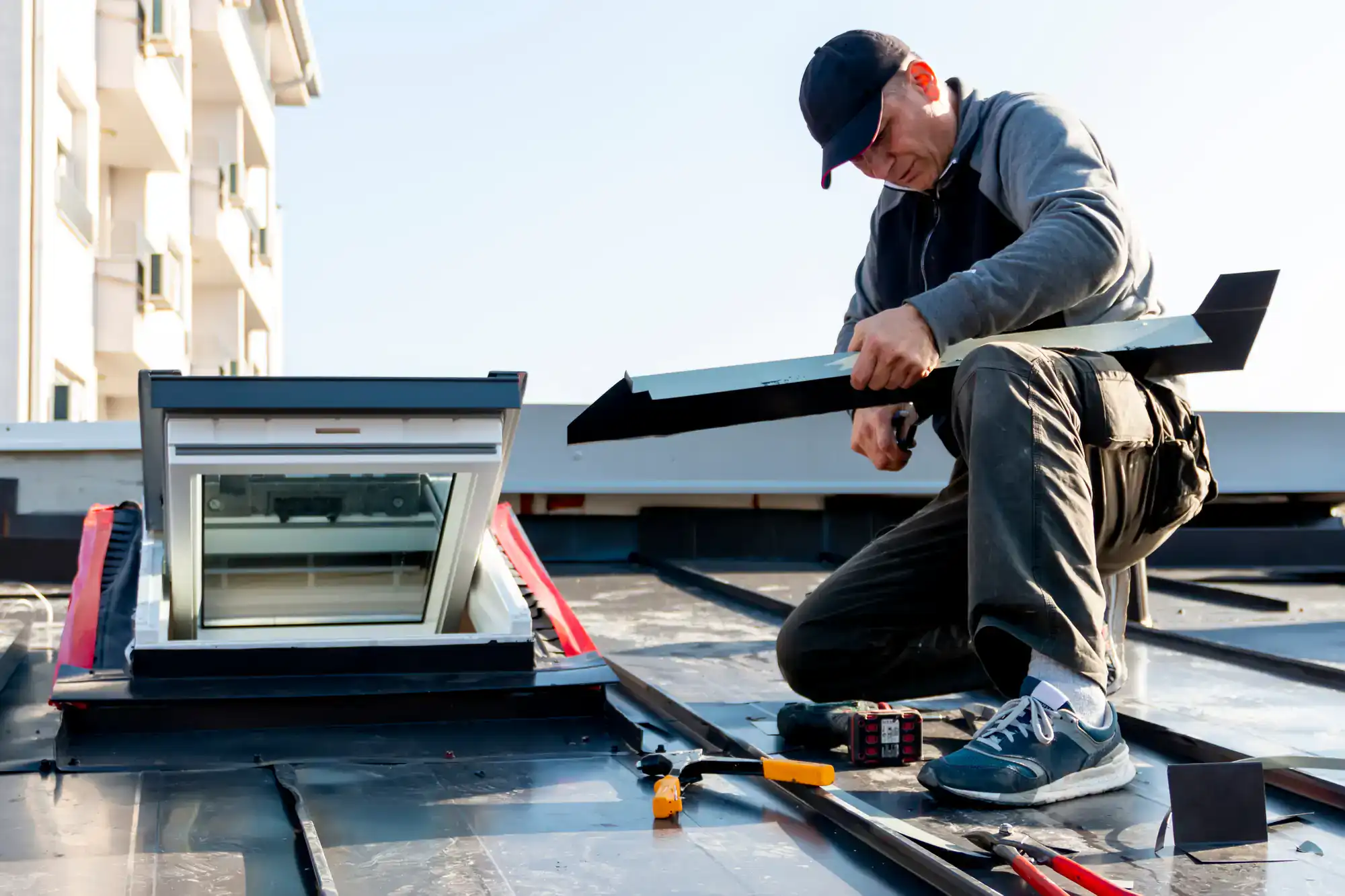 A person in work clothes and a cap kneels on a roof, installing or repairing a skylight window with various tools placed nearby on the roof surface. The sky is clear in the background.