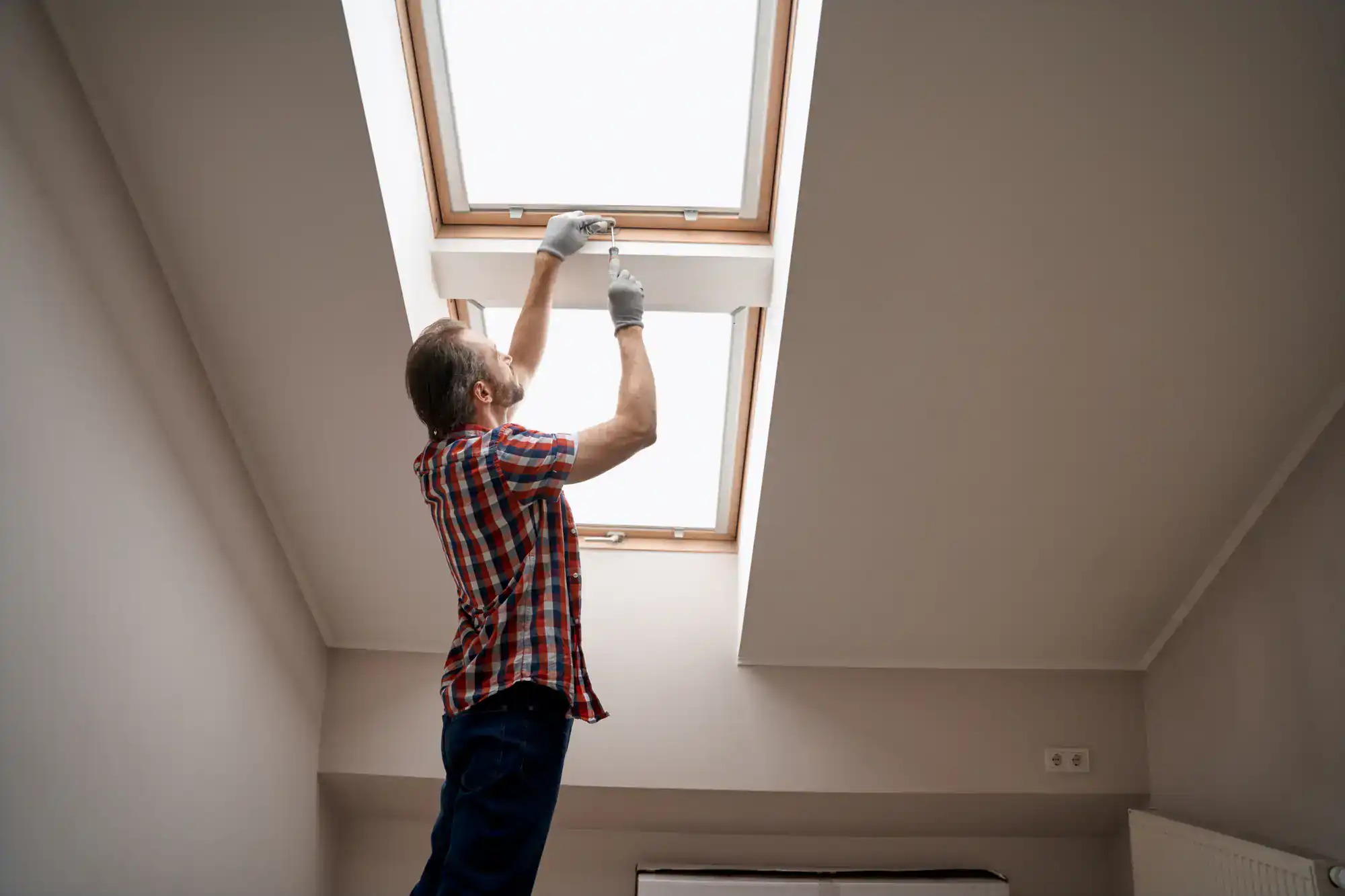 A man in a plaid shirt and gloves stands on a stool, cleaning or repairing a skylight window in a bright, modern attic room.