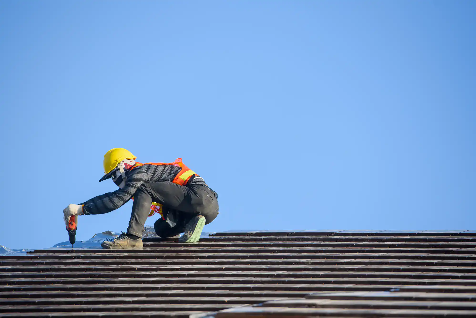 A construction worker wearing a yellow hard hat, safety vest, and gloves uses a power tool to work on a roof under a clear blue sky in Suffolk County, NY.
