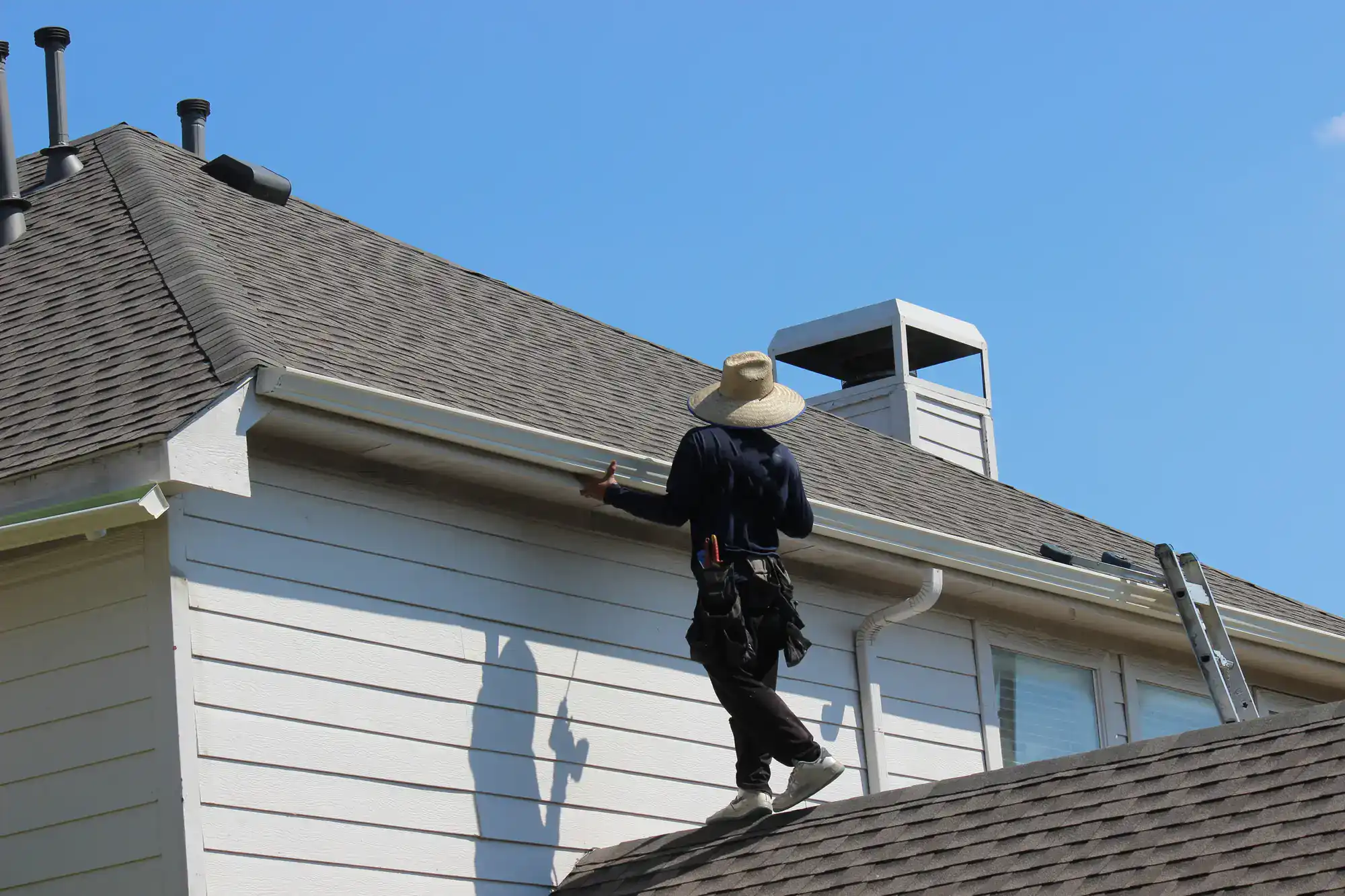 A person wearing a wide-brimmed hat and work gear stands on the roof of a house, inspecting or repairing the gutter under a clear blue sky. A ladder is positioned nearby against the roof.