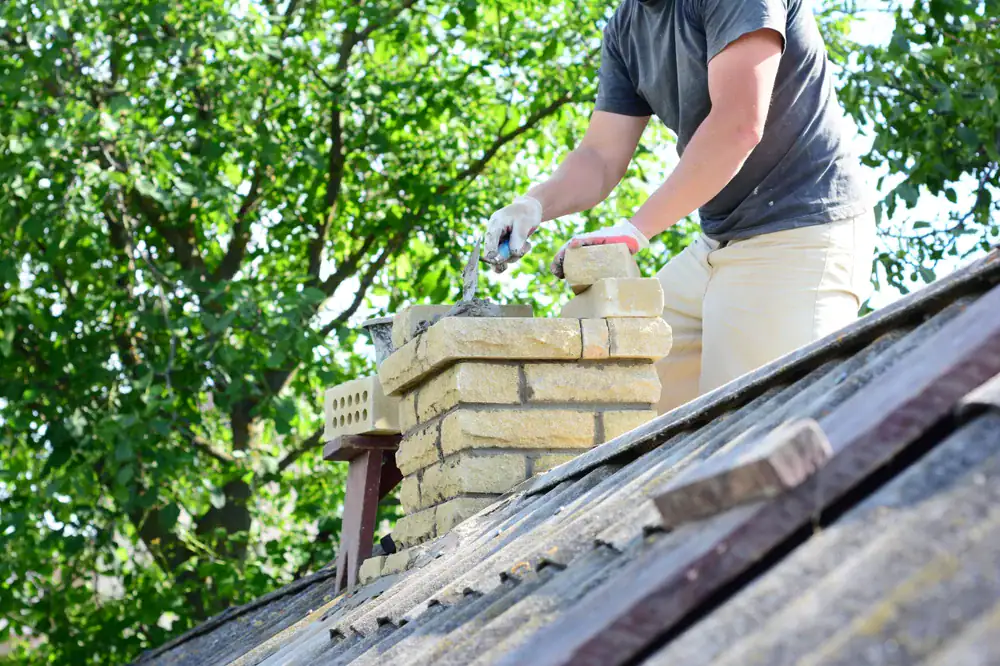 A person wearing gloves works on a brick chimney atop a slanted roof, using a trowel to lay bricks. Green leafy trees are visible in the background on a sunny day.
